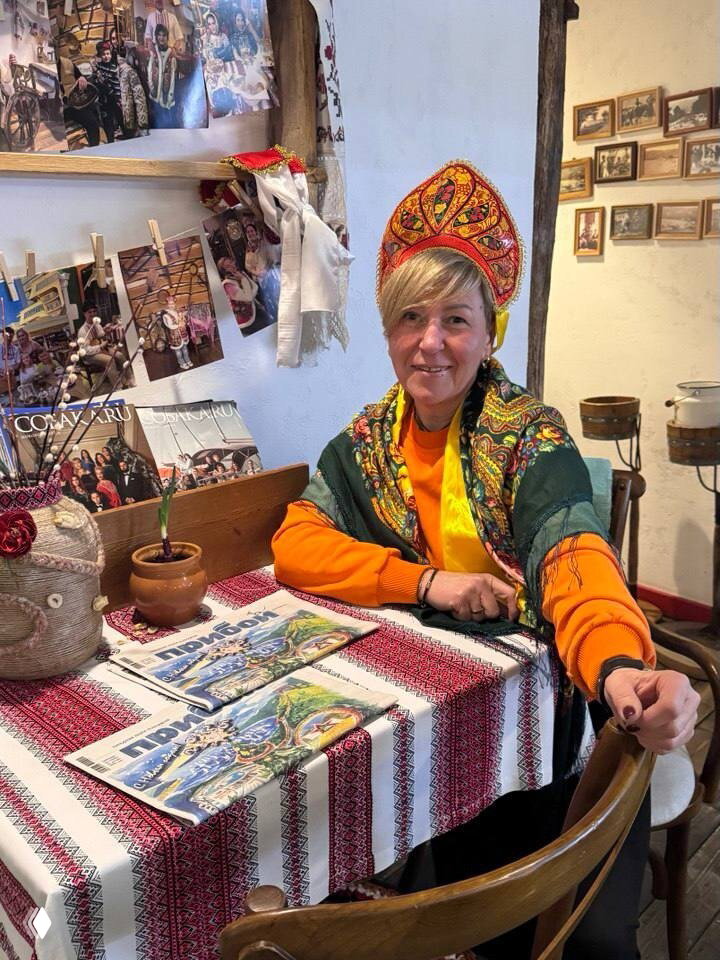 Женщина в традиционном русском кокошнике и платке сидит за столом с рукодельными сувенирами и журналами, интерьер музея/кафе.