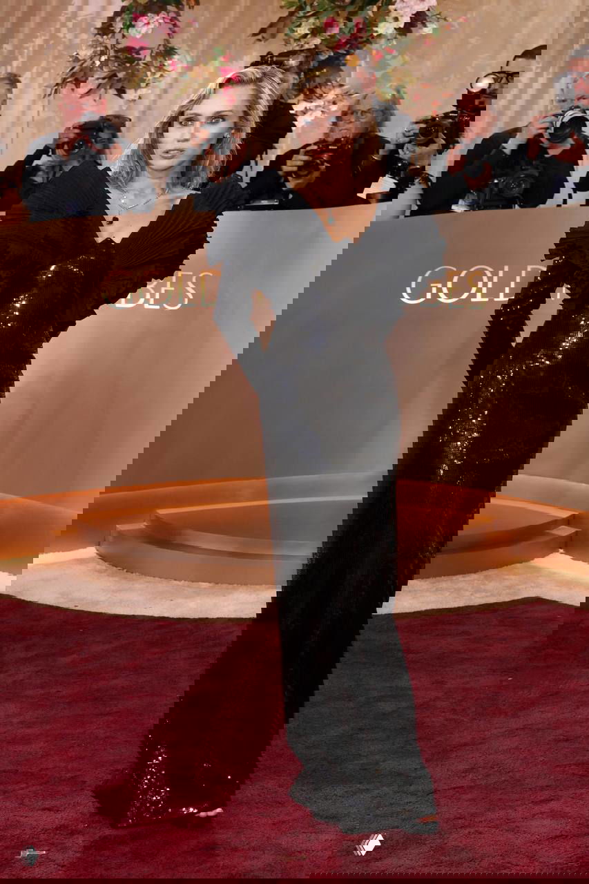 Дама в чёрном платье с выразительными рюшами и блёстками на красной дорожке, вспышки фотоаппаратов и папарацци позади неё.