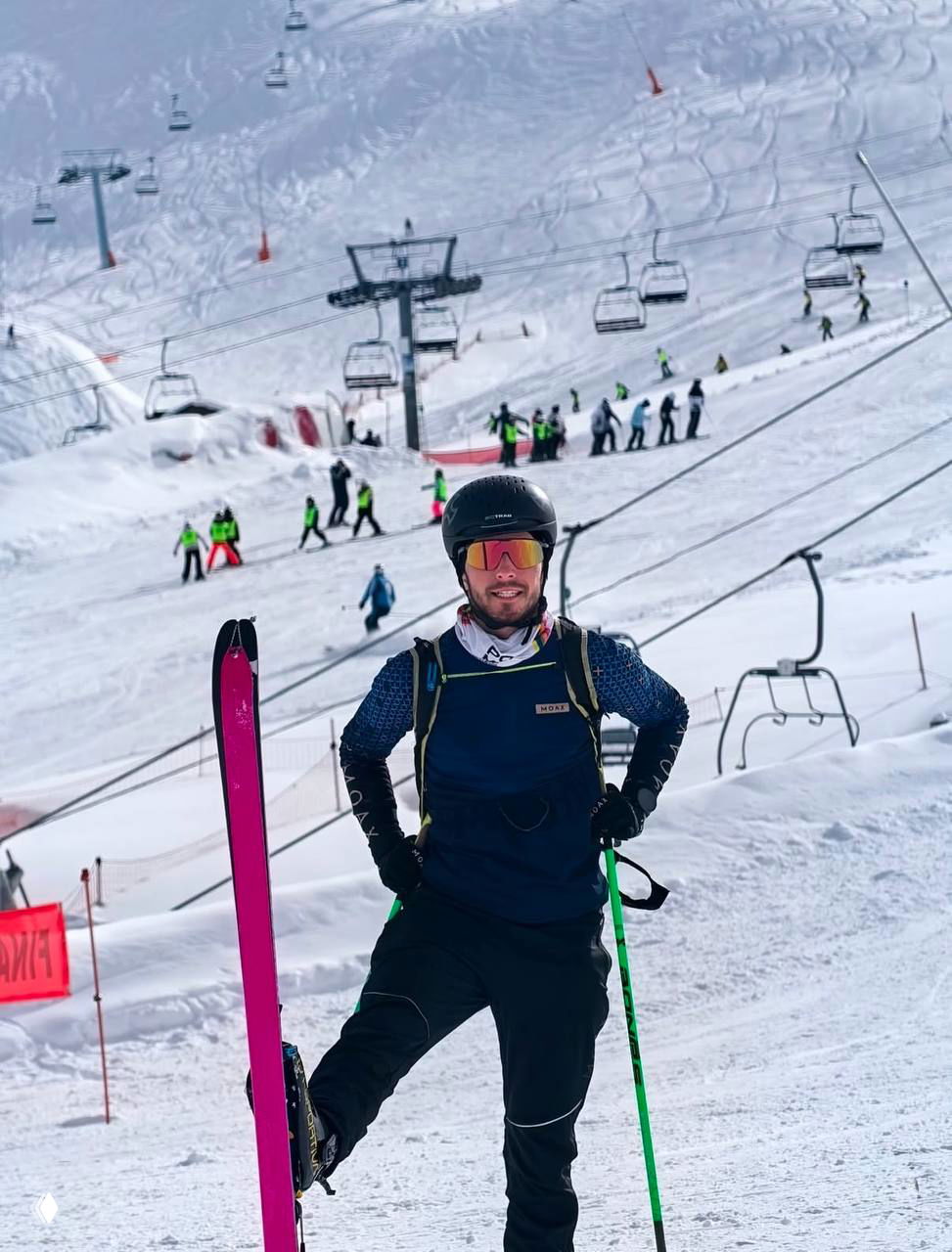 Атлет в защитном шлеме и очках стоит на заснеженном склоне рядом с яркой розовой лыжей; на заднем плане кресельные подъемники и другие лыжники на трассе.