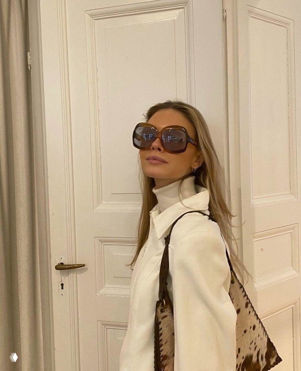 Девушка в больших квадратных солнцезащитных очках oversize и светлом образе в интерьере с белой дверью: элегантный ретро-шик и подчёркнутые пропорции аксессуара.