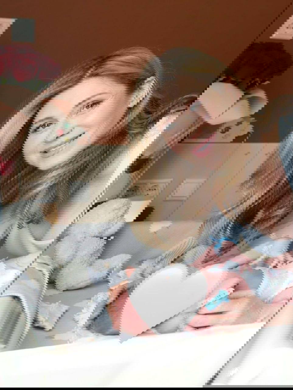 Актриса улыбается рядом с детской кроваткой, держит новорождённого; лицо младенца закрыто белым сердечком, на заднем плане виден букет.