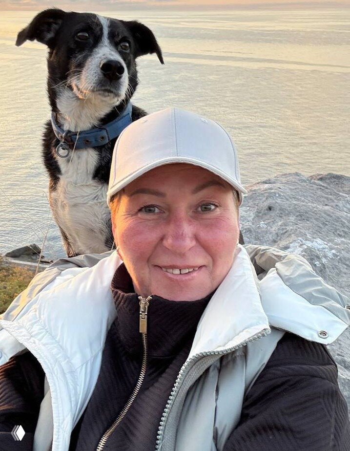 Селфи на фоне моря: женщина в светлой кепке улыбается, рядом виден её собака на берегу, закатное освещение и спокойная вода.