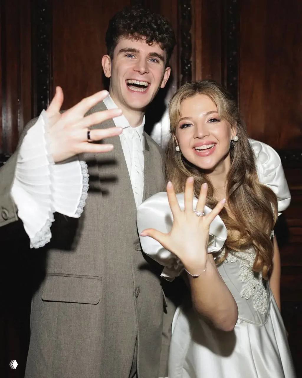 Аня и Артур Бабич улыбаются и демонстрируют руки с обручальными кольцами в праздничном портретном кадре; пара выглядит счастливой на фоне интерьера регистрации.