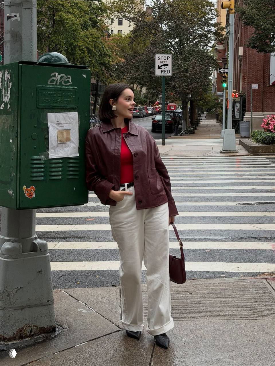 Девушка в бордовом жакете и молочном свитере с белыми брюками на пешеходном переходе в городском осеннем окружении.