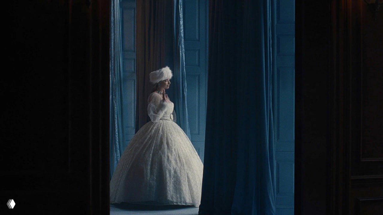 Марго Робби в том самом белом платье, снятая в полный рост у высоких окон зала; строгая интерьерная постановка подчёркивает готическую эстетику картины.