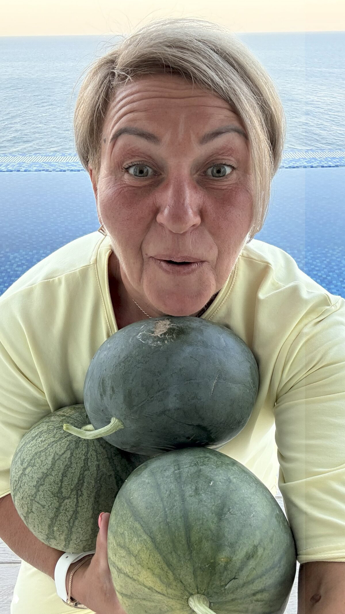 Крупное селфи женщины на фоне бассейна и моря, в руках несколько тёмно-зелёных арбузов — садовый урожай в кадре.