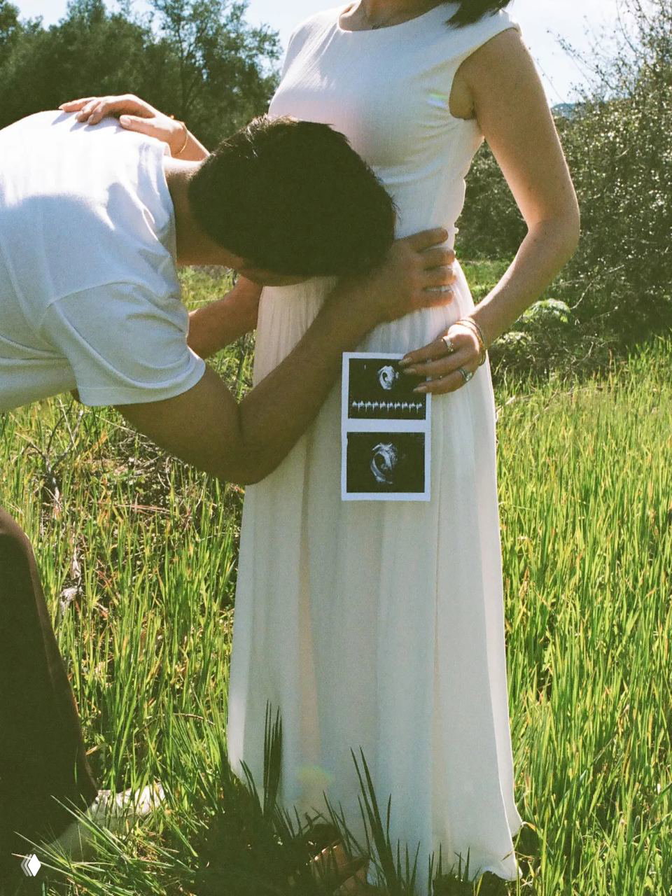 Мужчина целует живот беременной Тэй; на лёгком платье прикреплён УЗИ-снимок малыша, кадр снят на фоне травянистого поля.