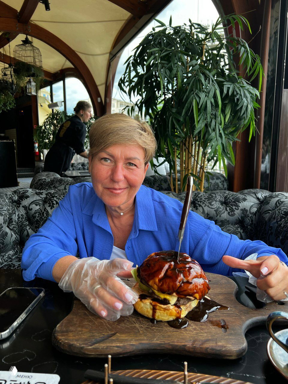 Портрет женщины в уютном кафе за столом; перед ней большой бургер на деревянной доске, атмосфера гастрономического снимка.
