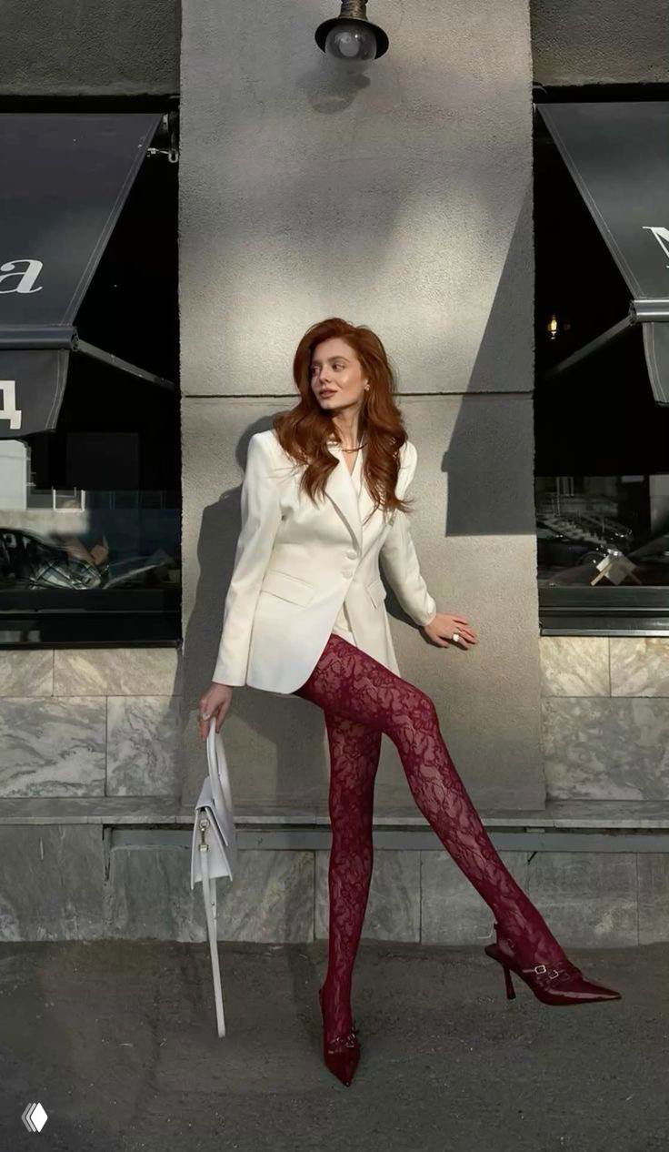 Модель в белом пиджаке и кружевных бордовых колготках на фоне фасада здания, высокий каблук и элегантная поза.