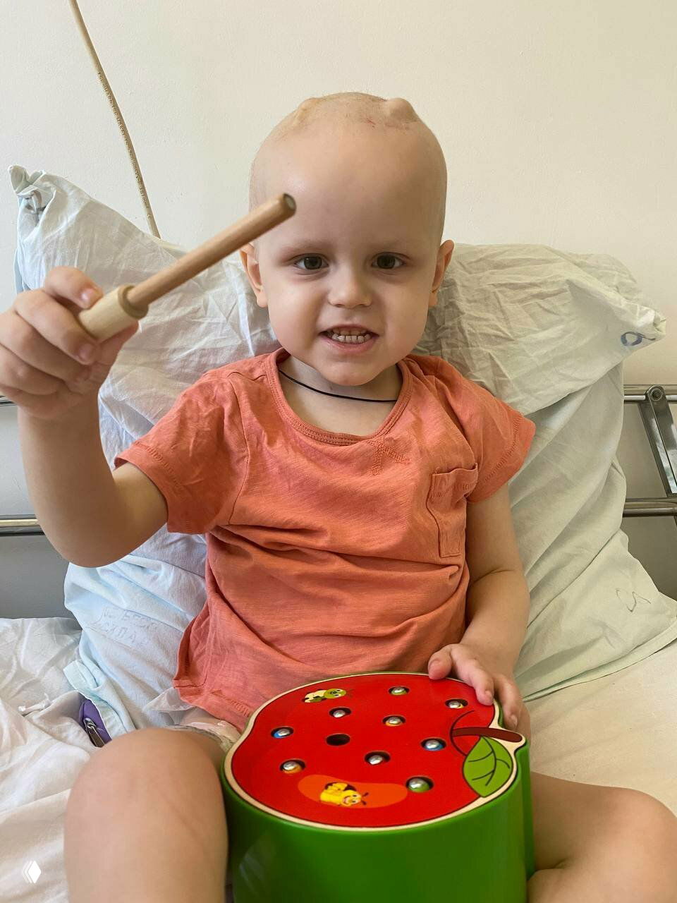 Маленький ребёнок с облысевшей головой в больничной палате играет деревянной игрушкой, сидя на кровати во время лечения.