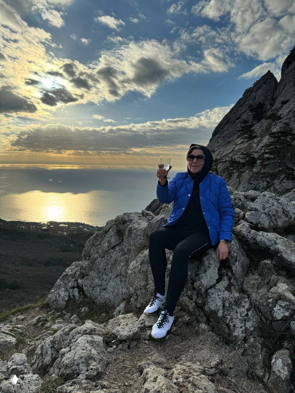 Женщина в куртке сидит на вершине горы с бокалом, на заднем плане море и закат — кадр походной и пейзажной тематики.