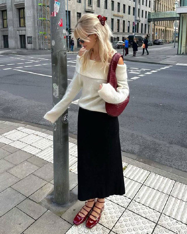 Блондинка в молочном свитере с открытыми плечами и длинной черной юбкой, бордовая сумка в тон обуви на городском тротуаре.