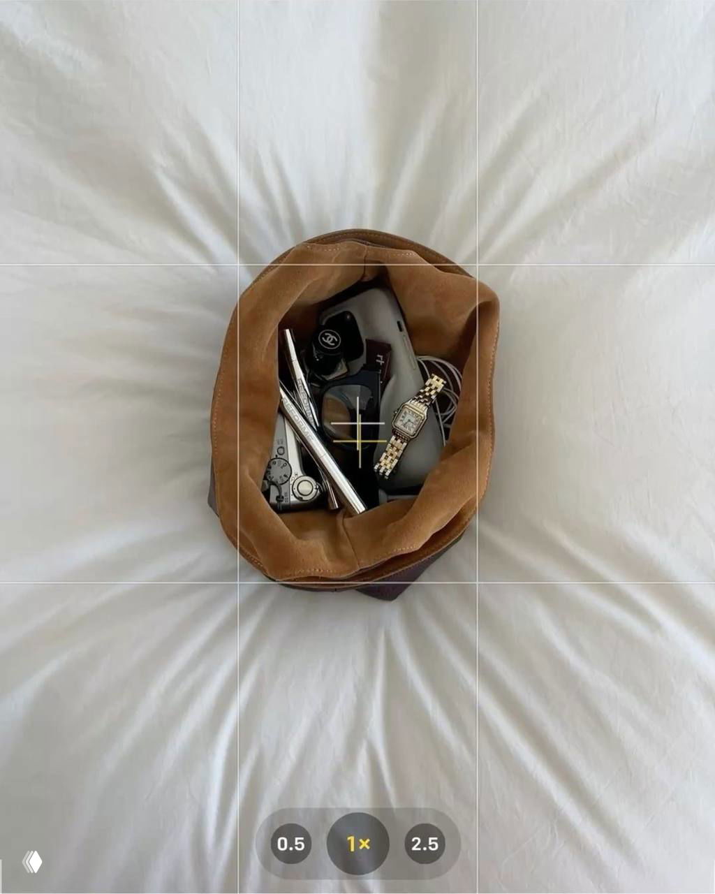 Вид сверху на открытый кошелёк или сумку на белом постельном фоне с аксессуарами: часы, очки, косметические предметы и камера.