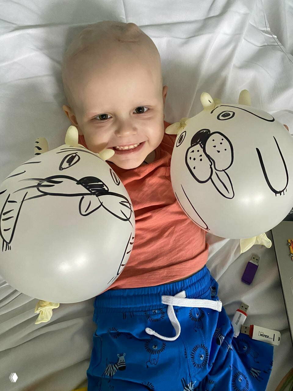 Улыбающийся ребёнок в больничной кровати держит два белых шарика с нарисованными мордами — светлая сцена благотворительного сбора.