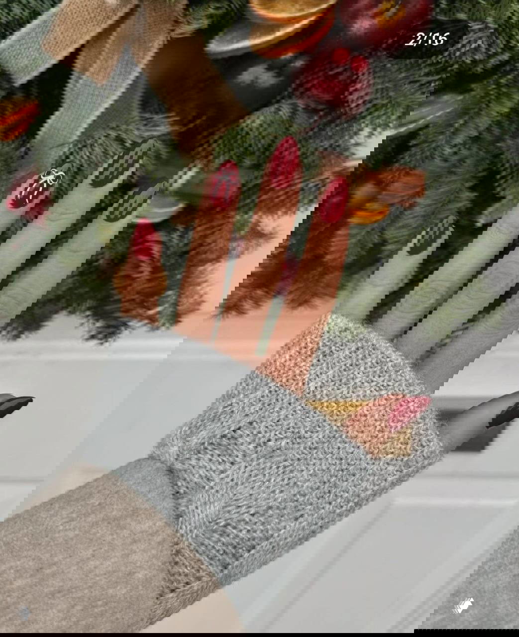 Рука с длинными овальными ногтями в глубокем бордовом цвете и маленьким белым бантиком на одном из ногтей, фон — праздничная ёлочная зелень.