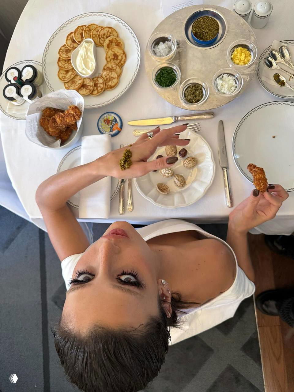 Вид сверху на сервированный стол с блинчиками, закусками и тарелками; женщина в кадре смотрит вверх — атмосфера утреннего приёма пищи.