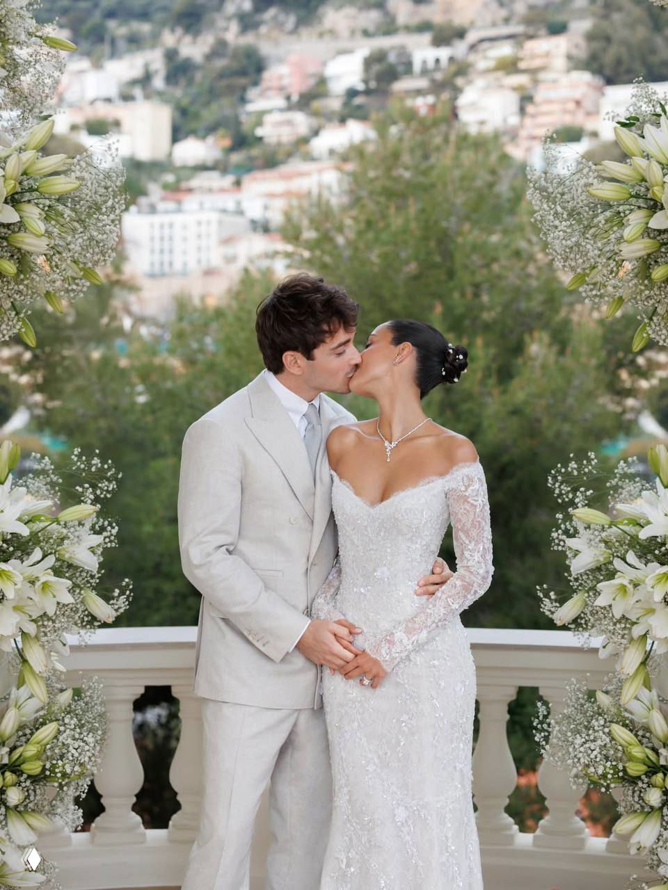 Молодые целуются на фоне зелёных насаждений и городской застройки; кадр украшен белыми цветами и создаёт романтическую атмосферу торжества.