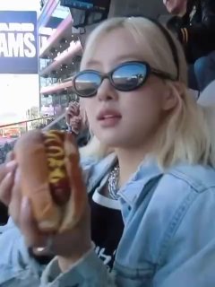 Женщина в джинсовой куртке и тёмных очках держит хот‑дог на фоне футбольного стадиона и экранов, видна толпа зрителей.