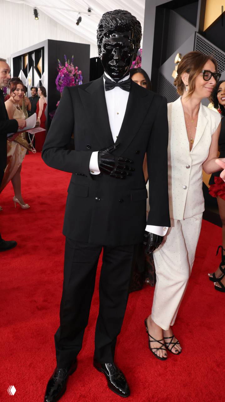 Мужчина с чёрным блестящим макияжем на лице в смокинге на красной дорожке «Грэмми», позирует возле фотозоны среди гостей, блестящая текстура кожи.