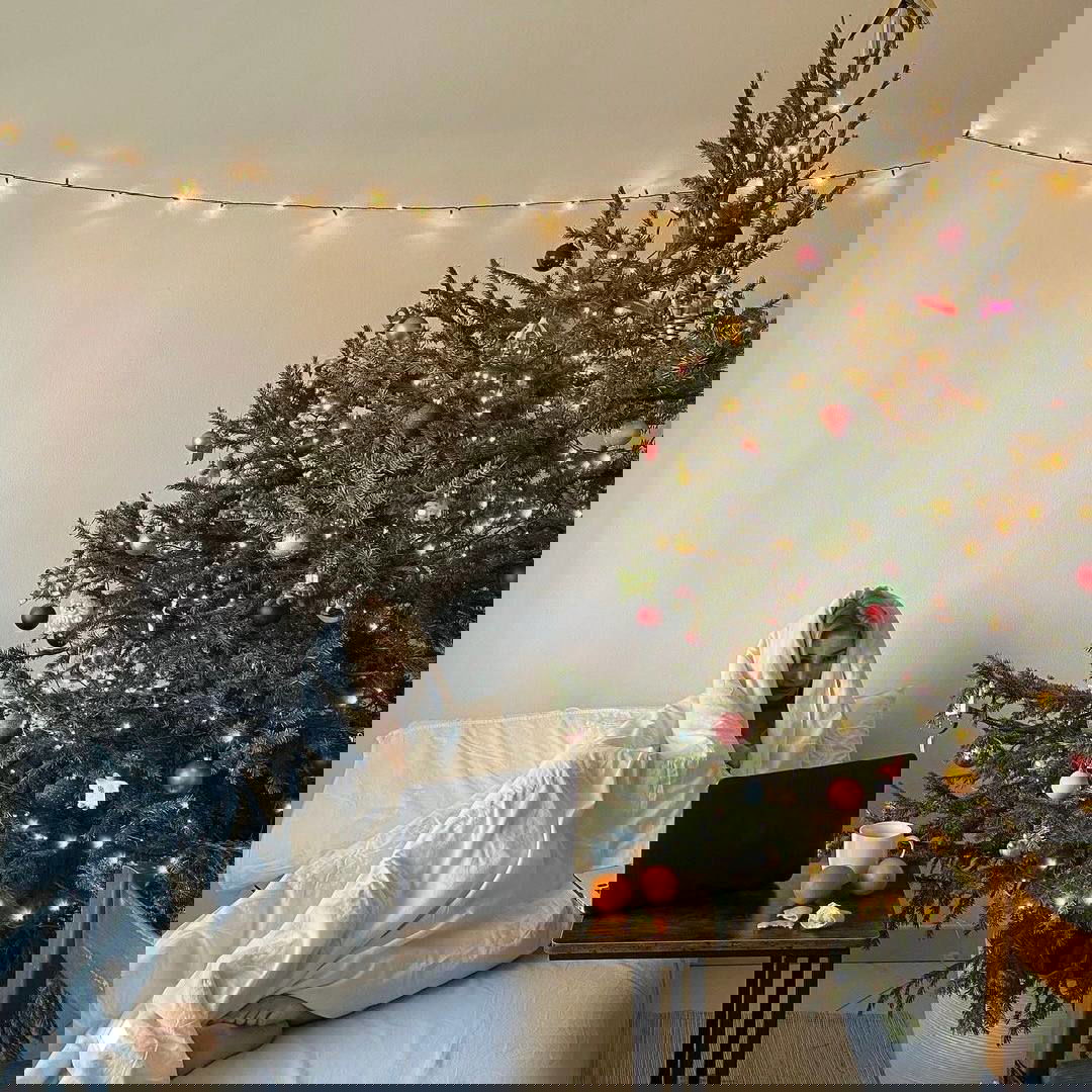 Девушка завернута в одеяло рядом с украшенной ёлкой, ноутбук и чашка рядом — кадр передаёт тихую рождественскую уютность и домашний комфорт.