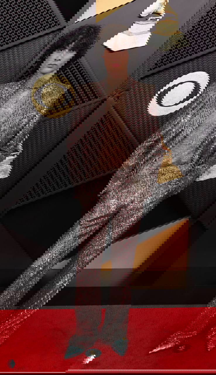 Мужчина в блестящем серебристом костюме и прозрачной кружевной рубашке на красной дорожке «Грэмми», полный рост на фоне логотипа, блестящие туфли.