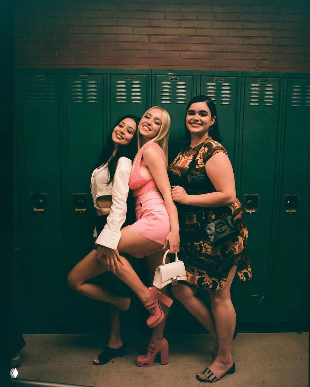Три актрисы позируют у школьных шкафчиков: яркая постановочная фотография с костюмами и стилизованным светом съёмочной площадки.