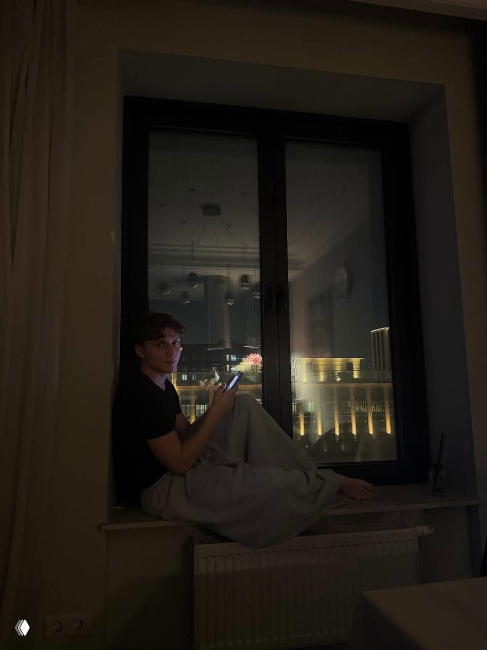 Молодой мужчина сидит на широком подоконнике вечером, держит телефон в руках, за окном ночные огни города и подсветка зданий, спокойная сцена.