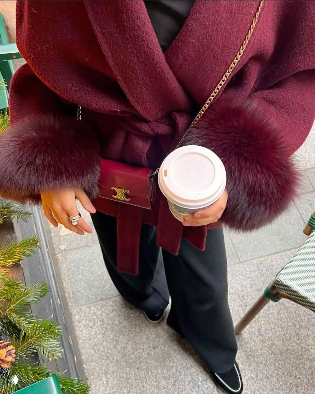 Крупный план бордового пальто с меховыми манжетами и маленькой лакированной сумкой; руки держат стакан кофе — городской уютный зимний лук.