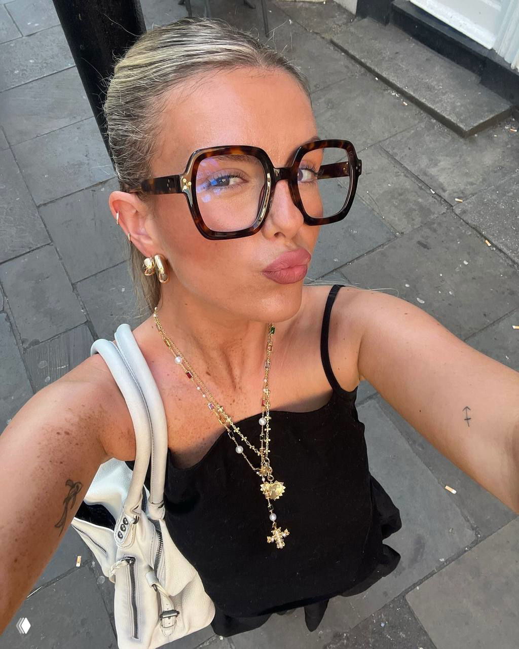 Селфи на тротуаре: женщина в полосатом кардигане и массивной сумкой демонстрирует средние по размеру очки с прозрачными линзами в повседневном образе.