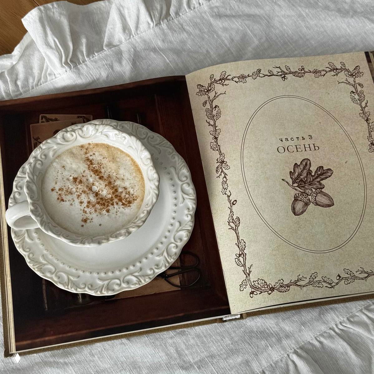 Открытая книга с разделом «ОСЕНЬ» и фарфоровая чашка с капучино на блюдце; атмосферный натюрморт, ассоциирующийся с чтением вкруг уютного утра.