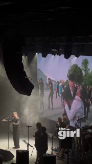 Широкий план сцены с ведущим исполнителем, оркестром и группой танцоров на фоне видеоряда и тёмной сценической подсветки