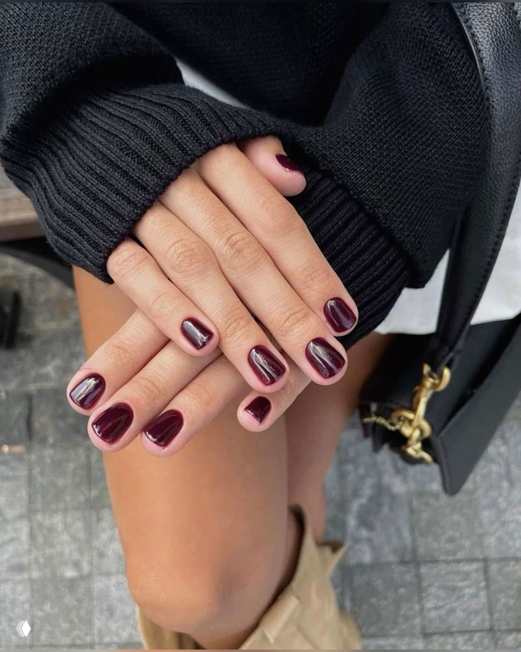 Короткий аккуратный бордовый маникюр на руках, лежащих на коленях; рядом видна сумка и бежевые сапоги — пример сочетания цвета ногтей с обувью и аксессуарами.