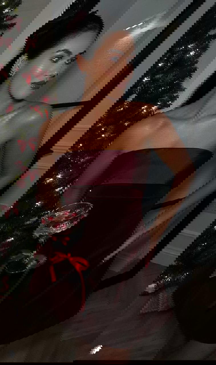 Женщина в коротком бордовом платье у украшенной ёлки держит праздничный бокал — гламурный вечерний образ с насыщенным винным цветом.