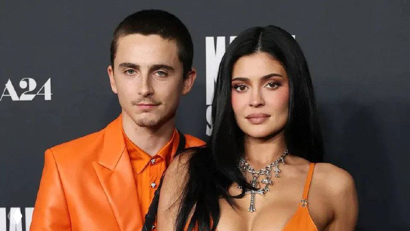 Двое людей в яркой одежде позируют на ковровой дорожке перед фотосессией; крупный план, логотипы и светящиеся баннеры на заднем плане