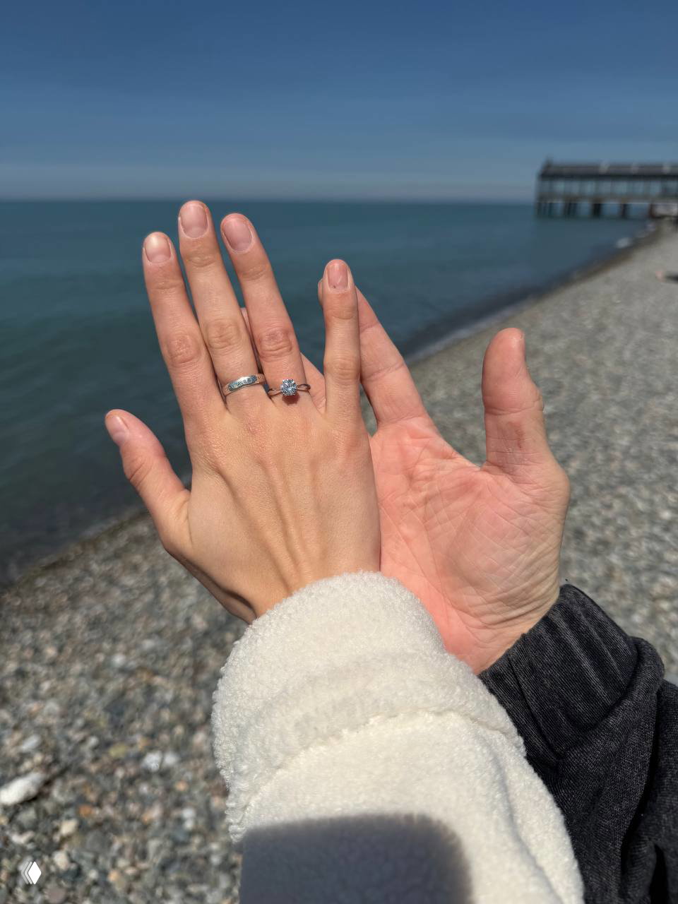 Крупный план рук с помолвочным кольцом и обручальным кольцом на фоне моря и пирса — подтверждение предложения на пляже.
