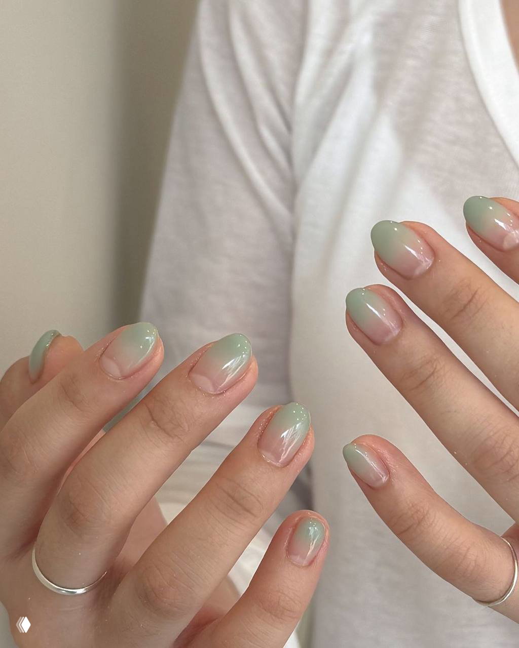 Детальный снимок двух рук с миндалевидной формой ногтей и мягким мятно‑полупрозрачным омбре; видны серебряные кольца и белая футболка на фоне.