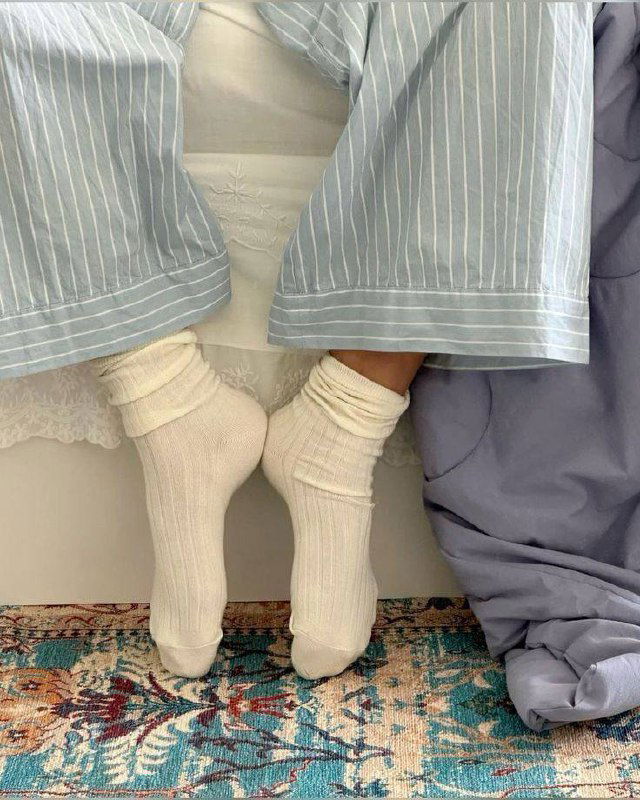 Кадр ног в светлых носках под пижамными брюками на кровати с узорчатым ковром и пастельным текстилем — уютная домашняя сцена.