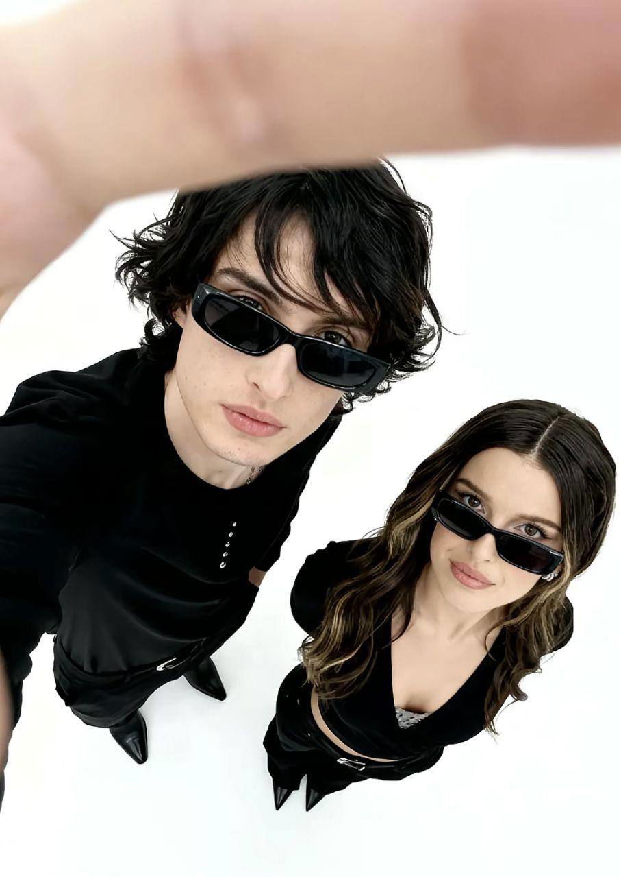 Два человека в чёрной одежде и тёмных очках сняты сверху на белом фоне — студийный кадр с минималистичной композицией.