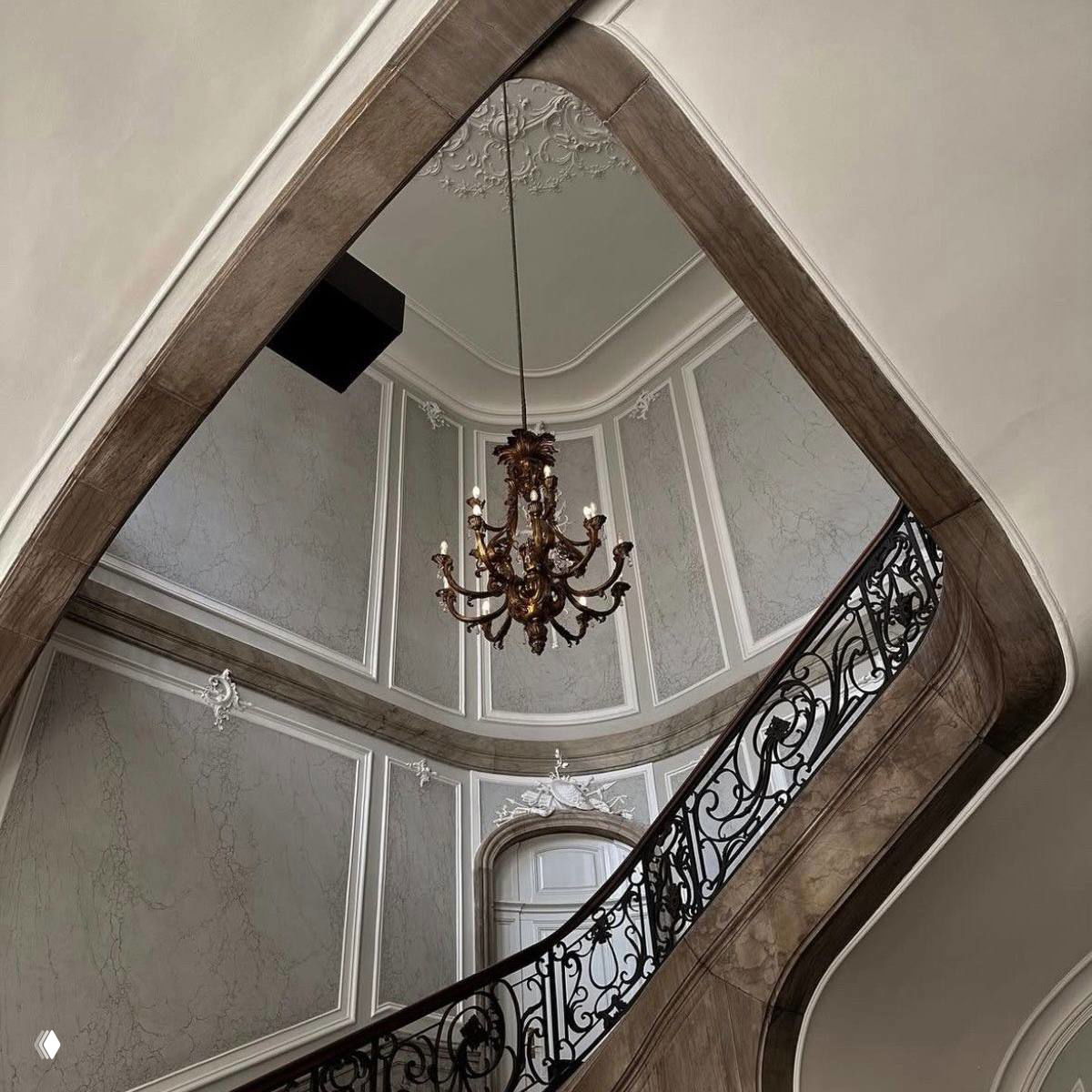 Винтажная лестница с коваными перилами и люстрой на фоне лепнины — интерьерная эстетика, навеянная классическими сюжетами и старинными домами.