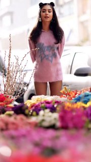 Яркий уличный кадр молодой женщины в розовом платье/свитере на фоне ряда цветочных киосков, передний план грубо размыт цветами.