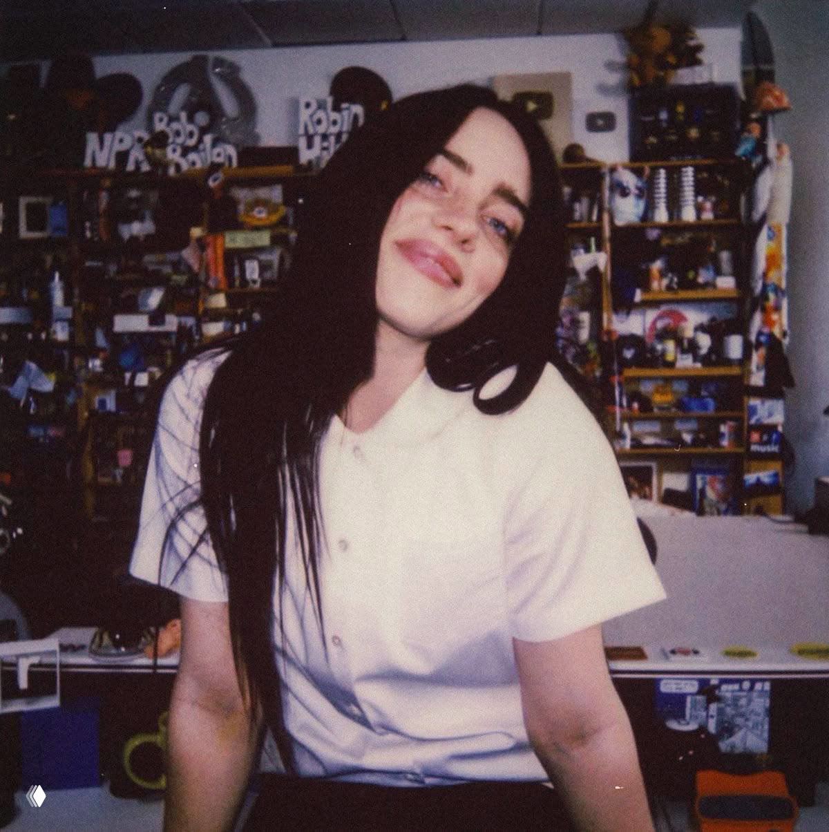 Портрет молодой женщины с тёмными длинными волосами, улыбается в уютной комнате с полками, плакатами и множеством предметов на заднем плане.