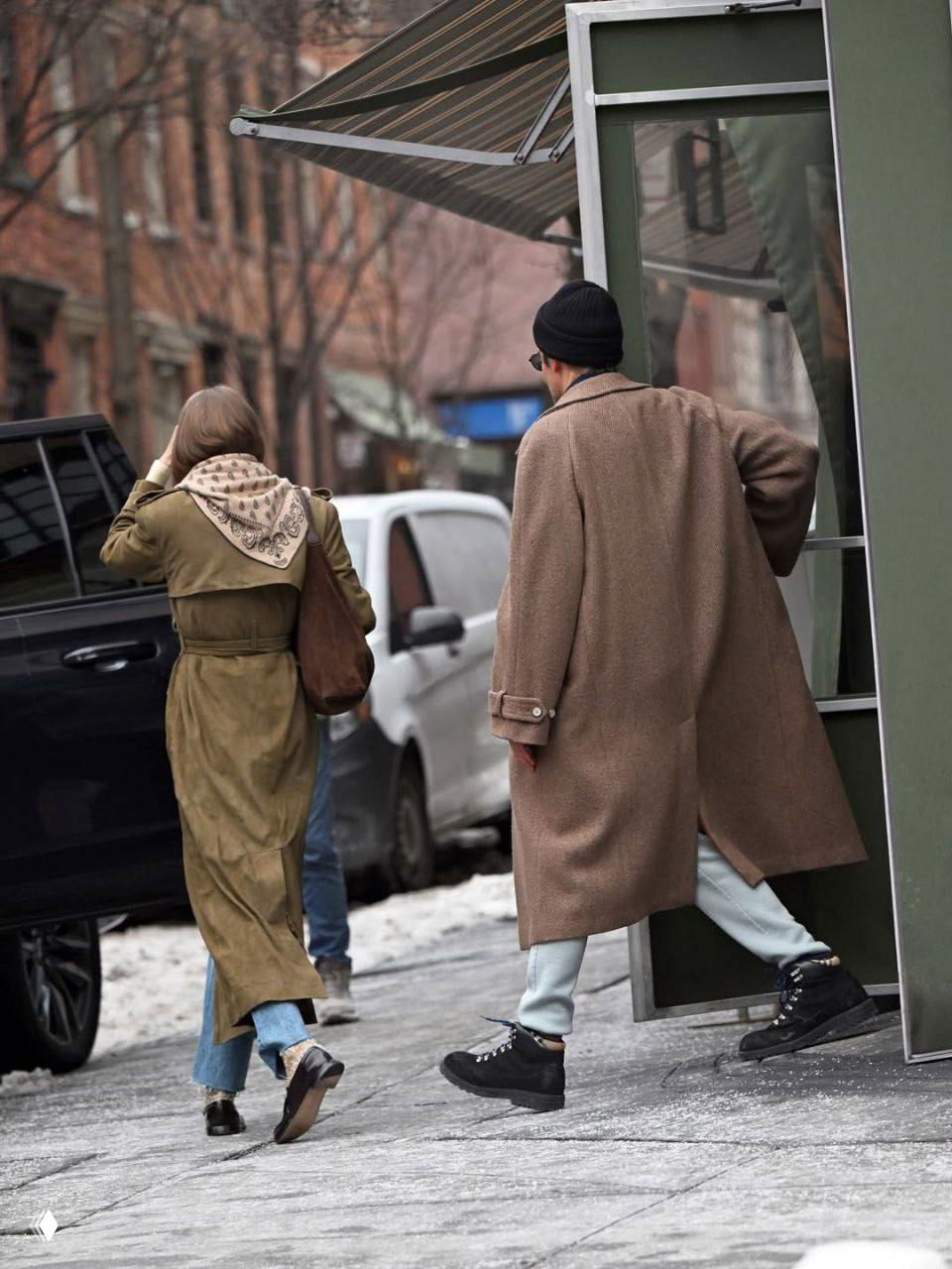 Кадр со спины: пара в пальто и замшевом тренче покидает тротуар в городе, заметны джинсы, ботинки и городской фон со снегом.