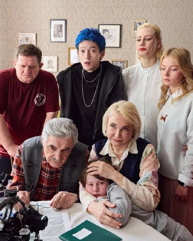 Группа актёров и персонажей в комнате: двое пожилых, молодой мужчина с синими волосами и девушка, семейный кадр со съёмочной обстановкой.