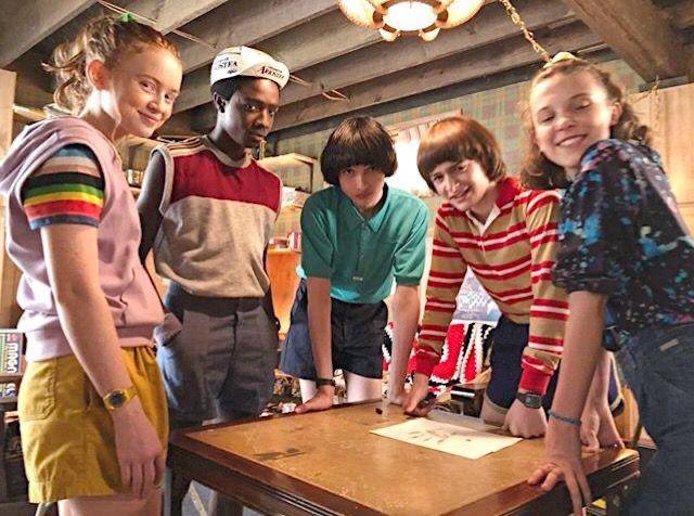 Съёмочная сцена: группа подростков за столом обсуждает план — бытовой кадр из сериала с реквизитом и дружеской атмосферой.