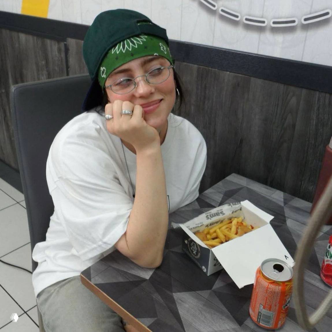 Женщина в кепке и бандане сидит за столом в кафе, перед ней коробка с картошкой фри и баночка газировки, расслабленная поза.
