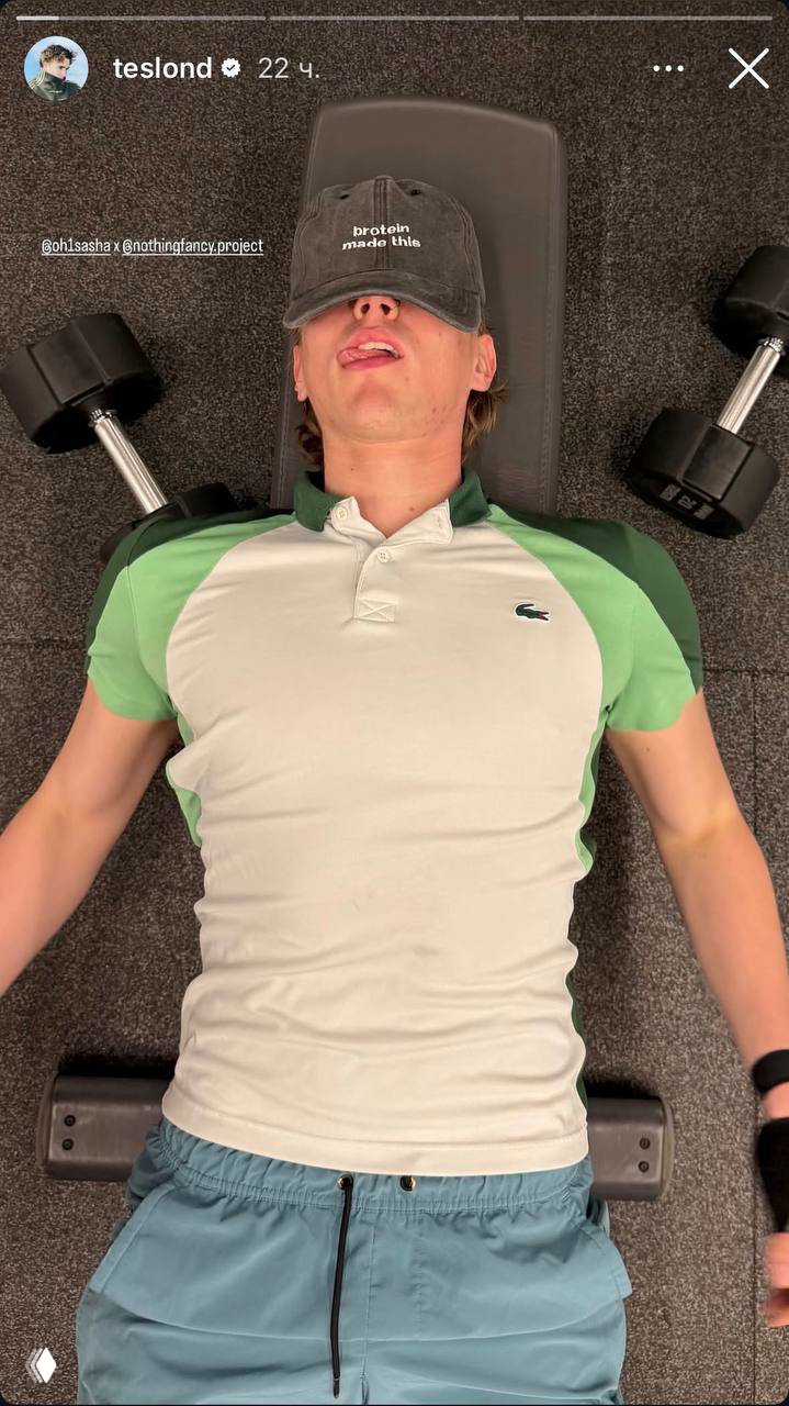Саша лежит на скамье в спортзале в кепке с надписью «protein made this», с гантелями по бокам — кадр после тренировки.