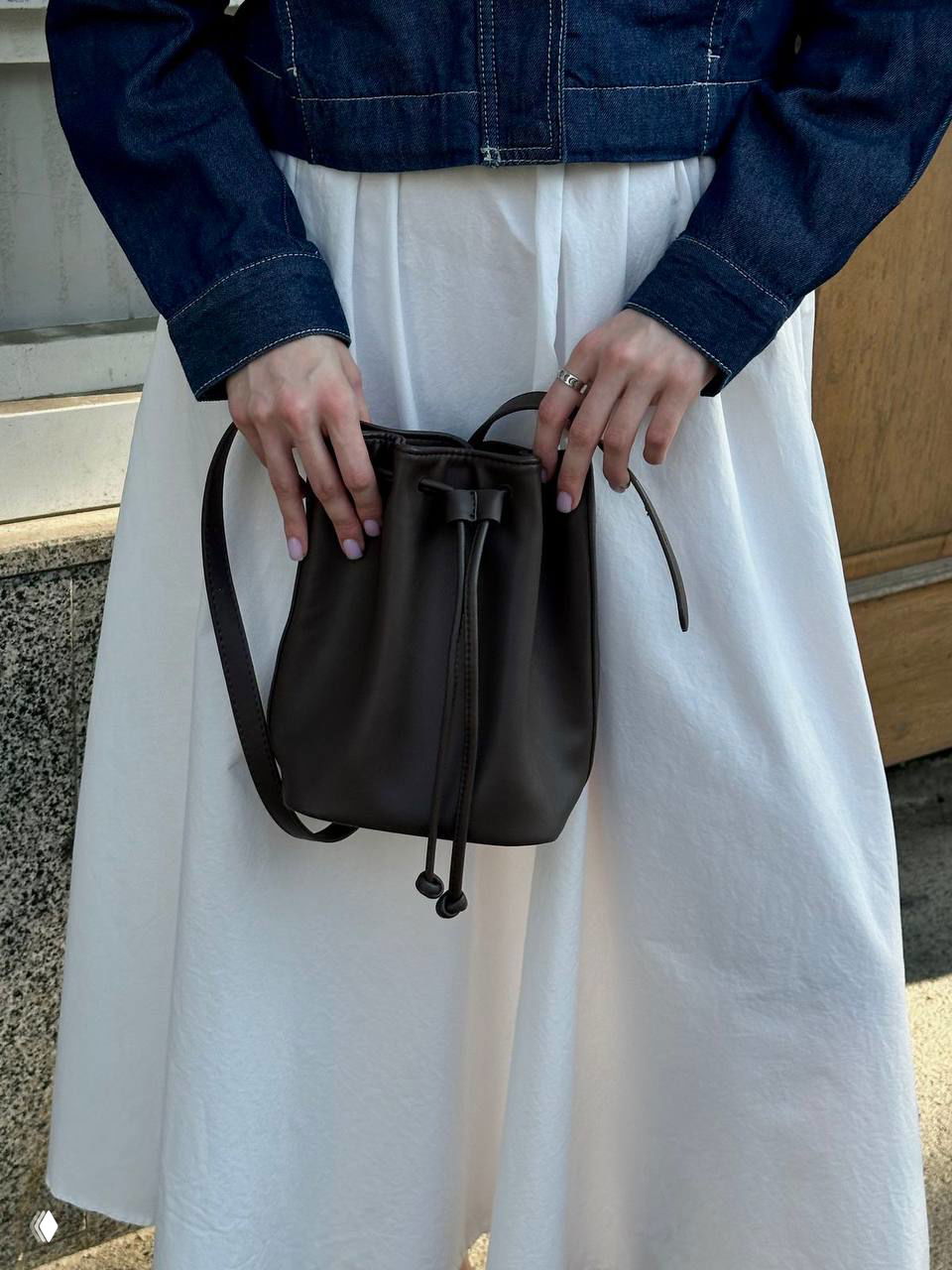 Небольшая кожаная сумка‑кисет тёмно‑коричневого цвета в руках у девушки на светлом платье, кадр подчёркивает шнурок и форму сумки.