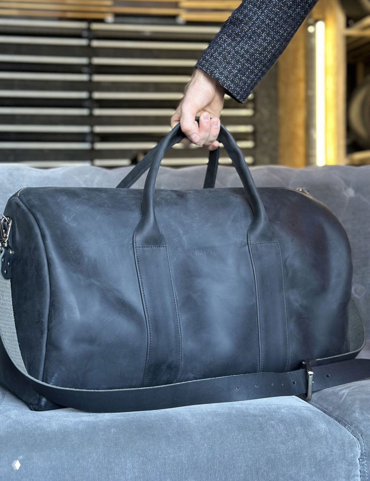 Чёрная дорожная кожаная сумка большого объёма на диване, ремень сумки лежит спереди, видна ручка и швы изделия ручной работы.