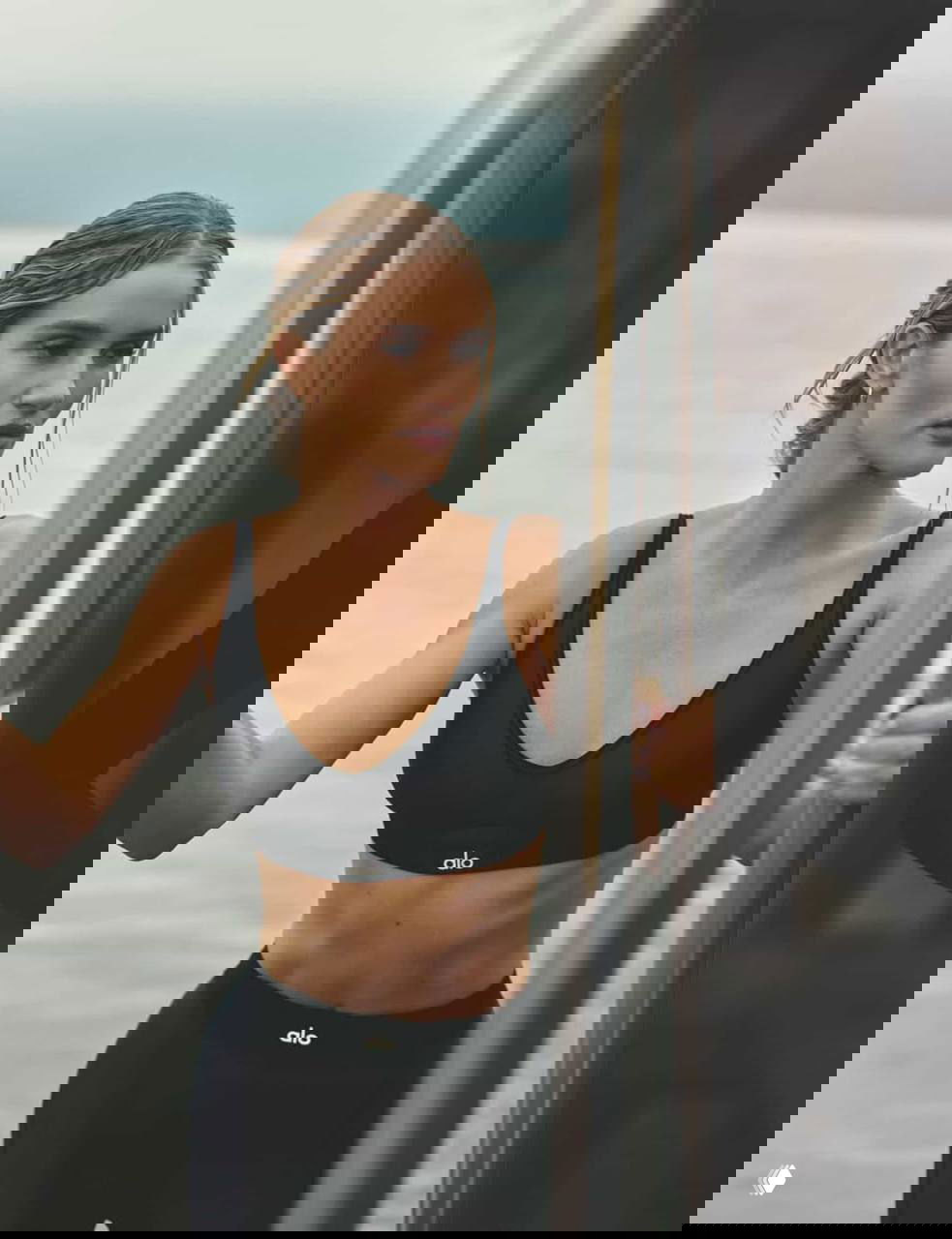 Модель в чёрном спортивном топе и лосинах у воды, крупный план туловища и лица, естественный макияж и спокойная поза на причале.