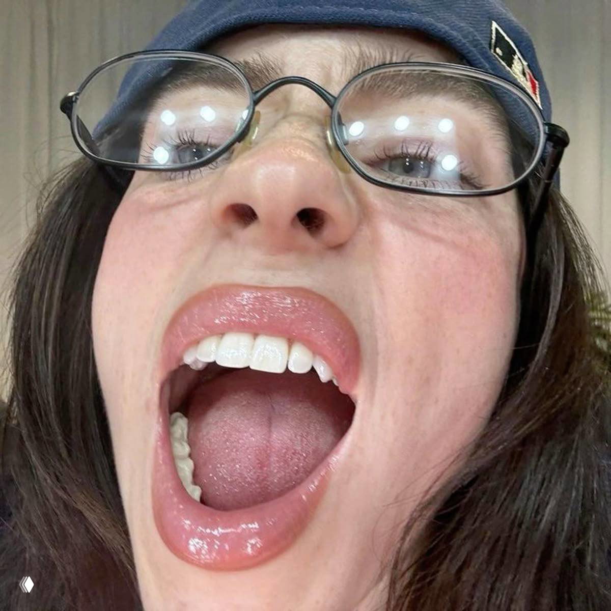 Крупный эмоциональный план женщины в очках с приоткрытым ртом, видны зубы и язык, на очках отражается несколько точечных источников света.